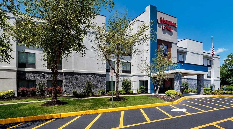 Exterior view of a Hampton Inn hotel with handicap parking space in the foreground, trees, landscaping, and a clear blue sky.