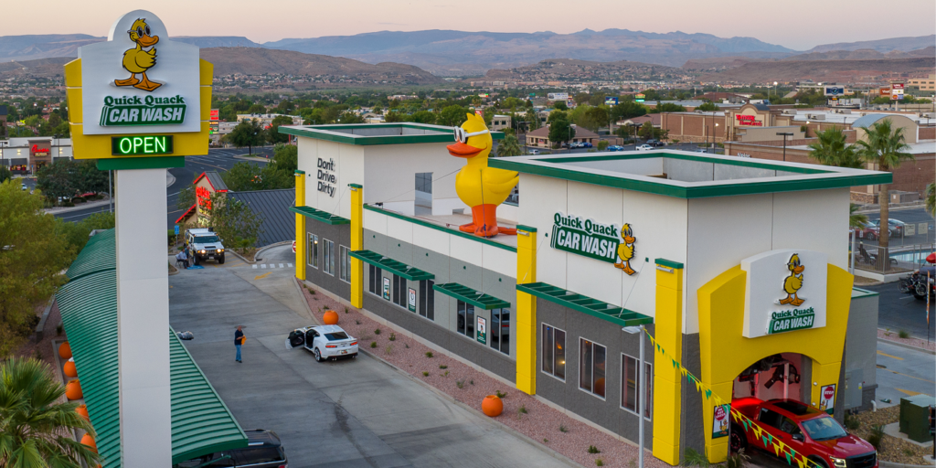 Exterior view of Quick Quack Car Wash with a large duck mascot on the roof, colorful signage, and a drive-through area with a few cars, in a commercial area with mountains in the background.