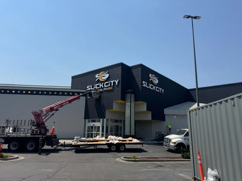 Construction workers installing a sign at Slick City Bowling & Arcade. A crane truck is parked in front, and a gray construction fence surrounds the site.