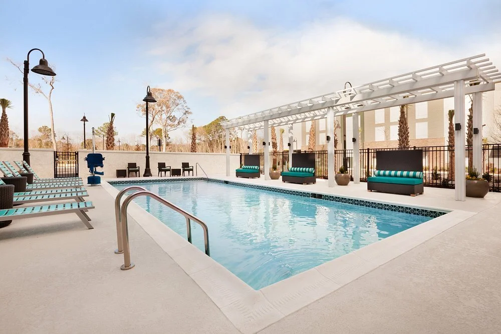 Empty outdoor swimming pool with lounge chairs, a shaded seating area, and decorative lamps, surrounded by a fence and trees under a partly cloudy sky.