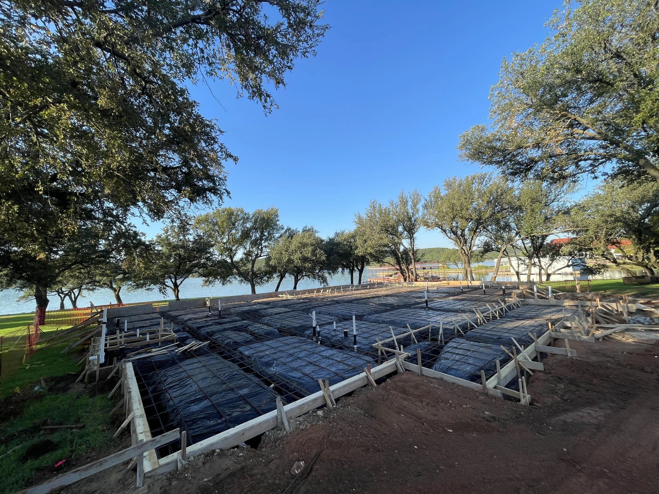 Construction site near a lake with trees and a blue sky. The concrete foundation is being prepared with wooden forms and rebar.