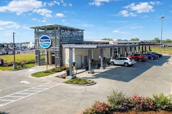 Exterior of a car wash facility with a drive-through area, several parked cars, and a sign indicating 'Car Wash' under a blue sky.