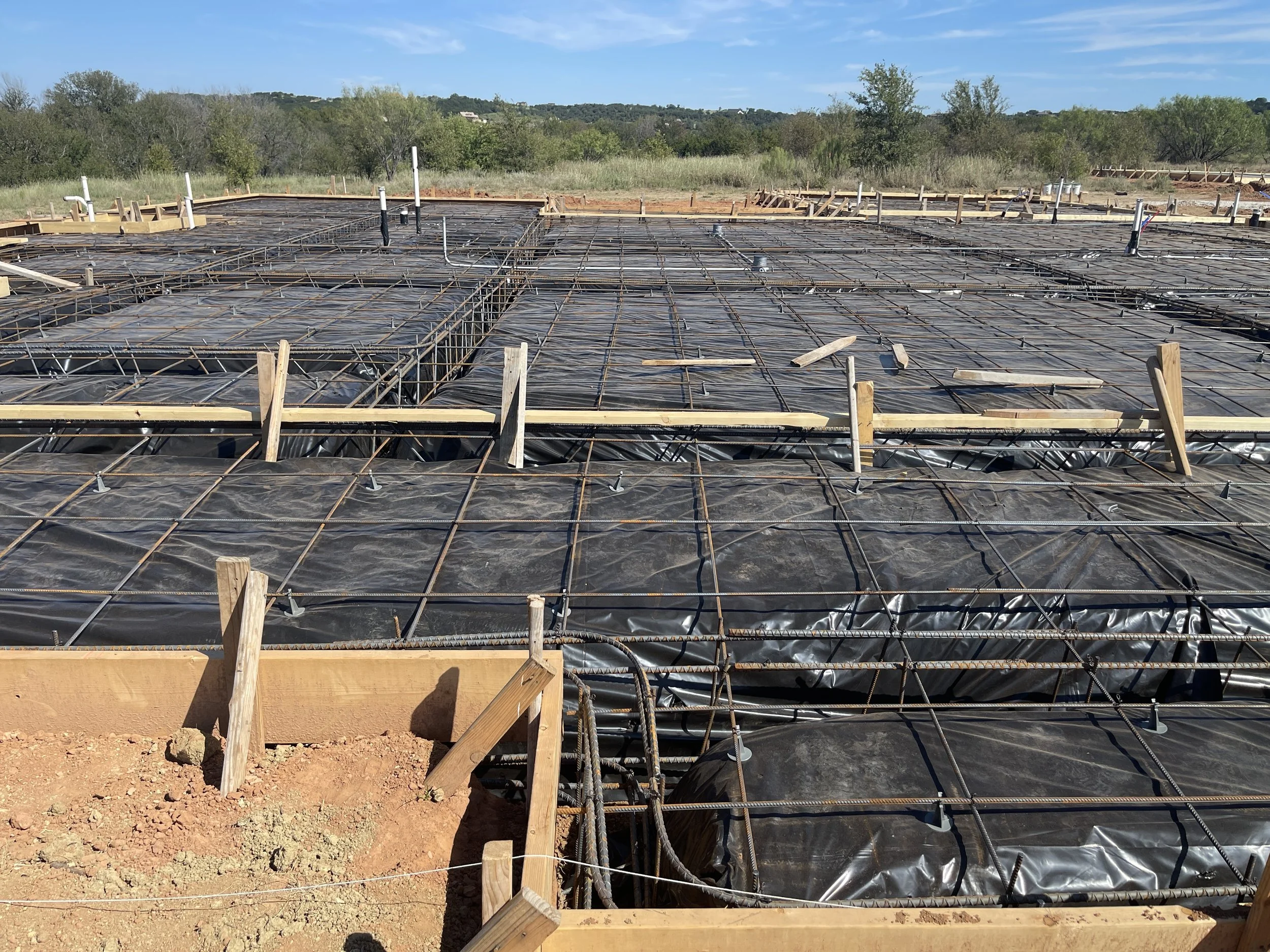Construction site with foundation framing and rebar for a building, set outdoors with trees and blue sky in the background.