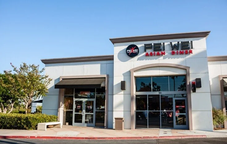 The exterior of PEI WE Asian Diner, a restaurant with a white facade, featuring a large glass entrance door with an 'Open' sign, and a logo with a black circle and red elements. There is greenery and a bench to the left, and the sky is clear.