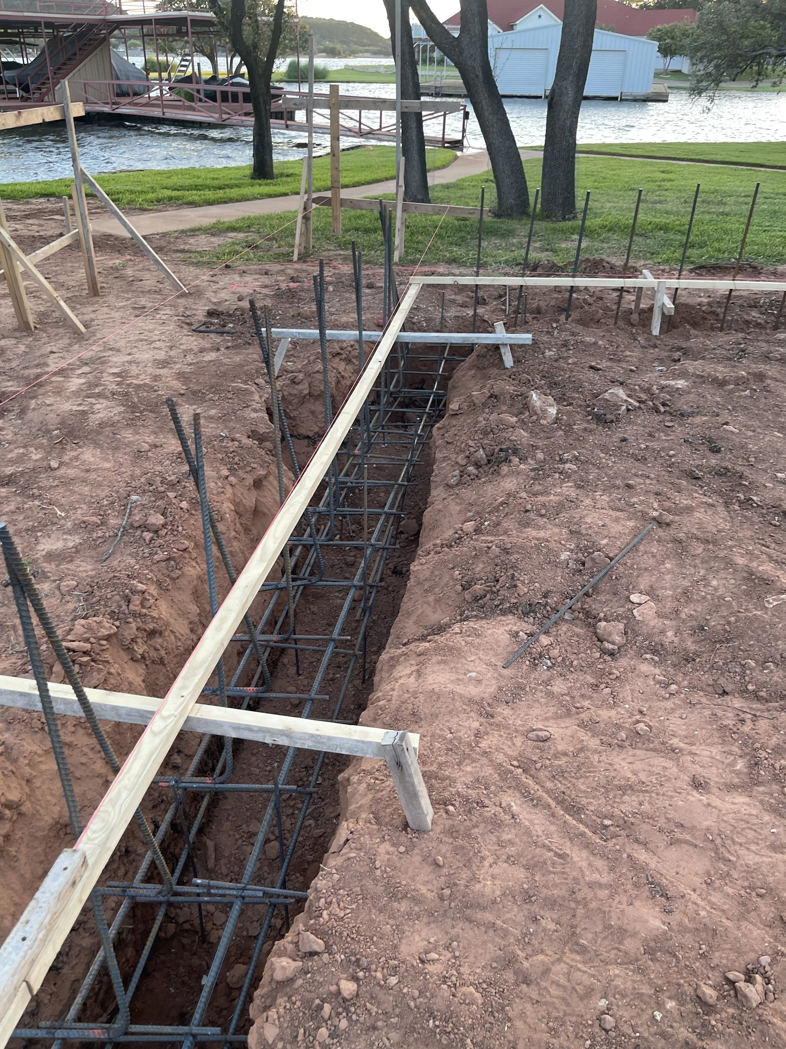 Construction site showing a foundation being prepared with rebar and wooden formwork near a park with trees, water, and buildings in the background.