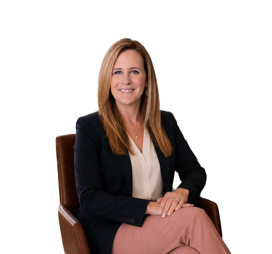 A woman with long red hair, smiling, sitting on a brown chair against a black background. She is wearing a black blazer, a light blouse, and pink pants.