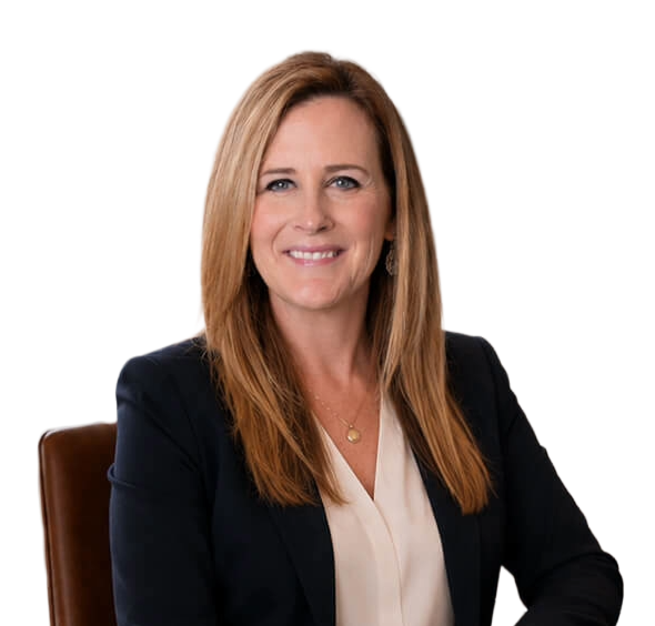 A woman with long red hair, wearing a black blazer and white blouse, seated at a table, smiling at the camera.