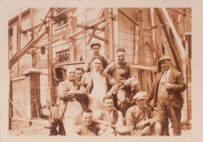 A group of men and women posing in front of a wooden construction site, with some standing and others sitting, likely from a historical period.