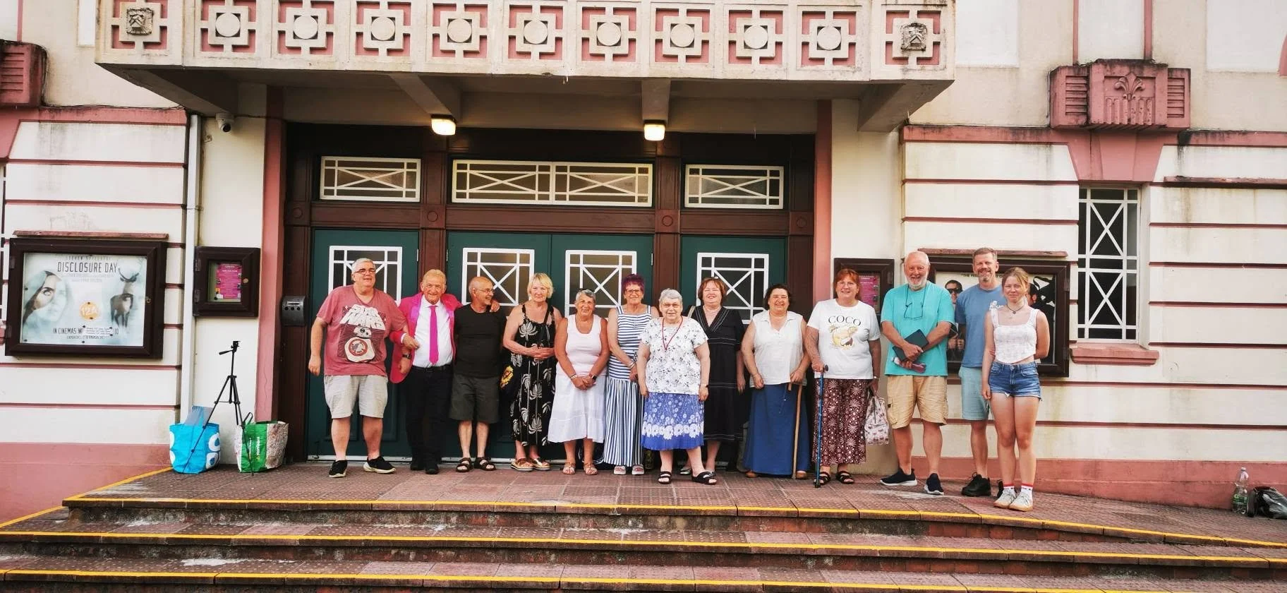 A group of 14 people standing on steps in front of a building with decorative windows and doors, smiling for the photo.