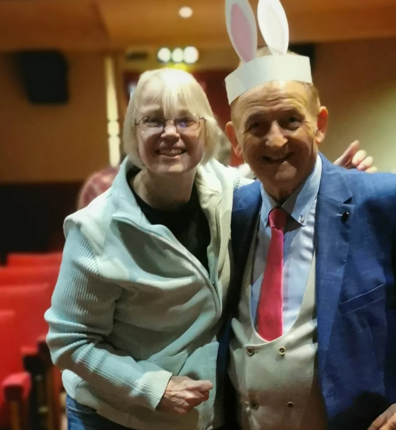 A woman and a man are smiling and posing for a photo in a warmly lit room. The man is wearing a bunny ears headband, a blue blazer, a white shirt, and a pink tie.