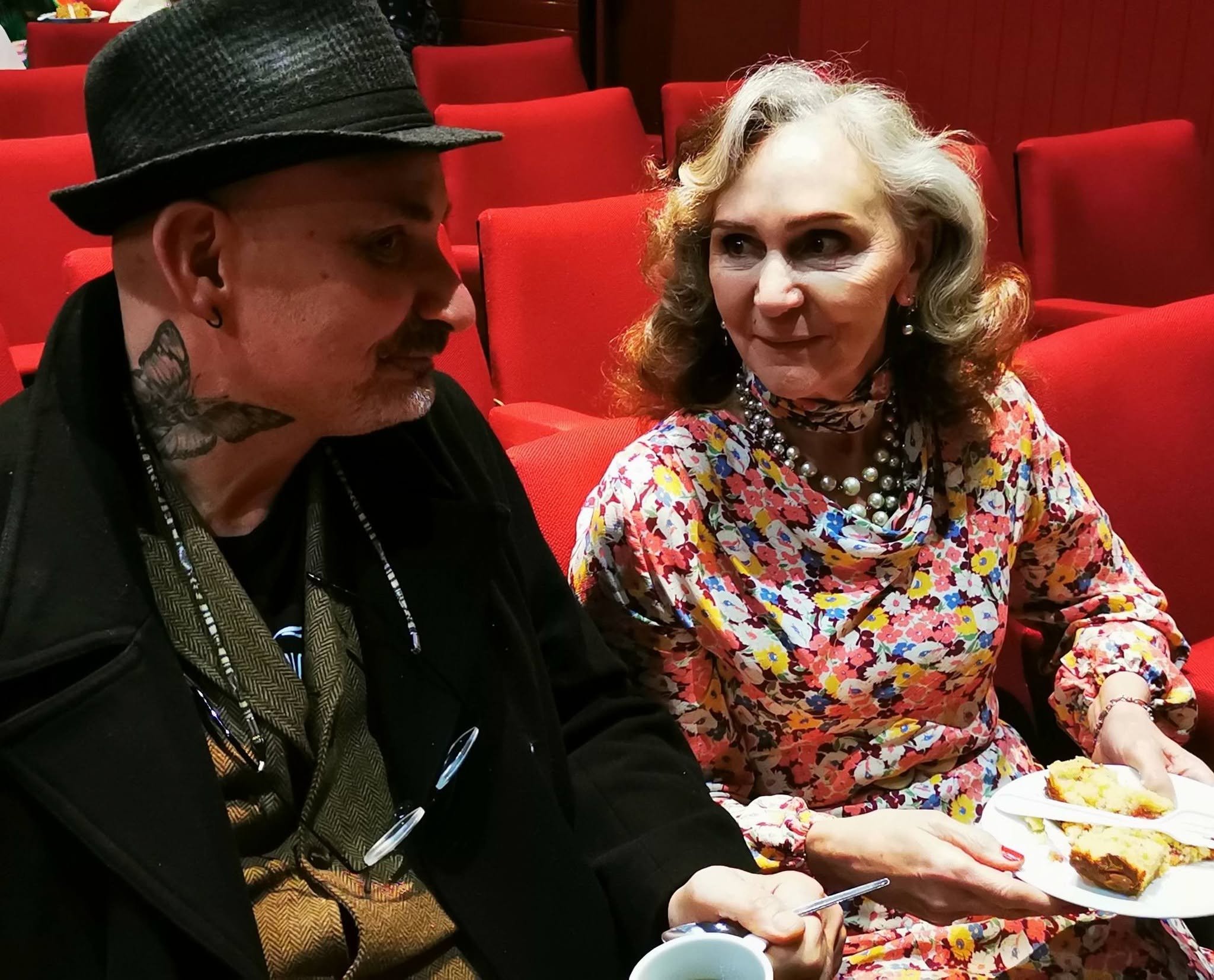 A man with tattoos and a woman with earrings and a pearl necklace sitting in a theater with red seats, sharing a plate of cake or pastry.