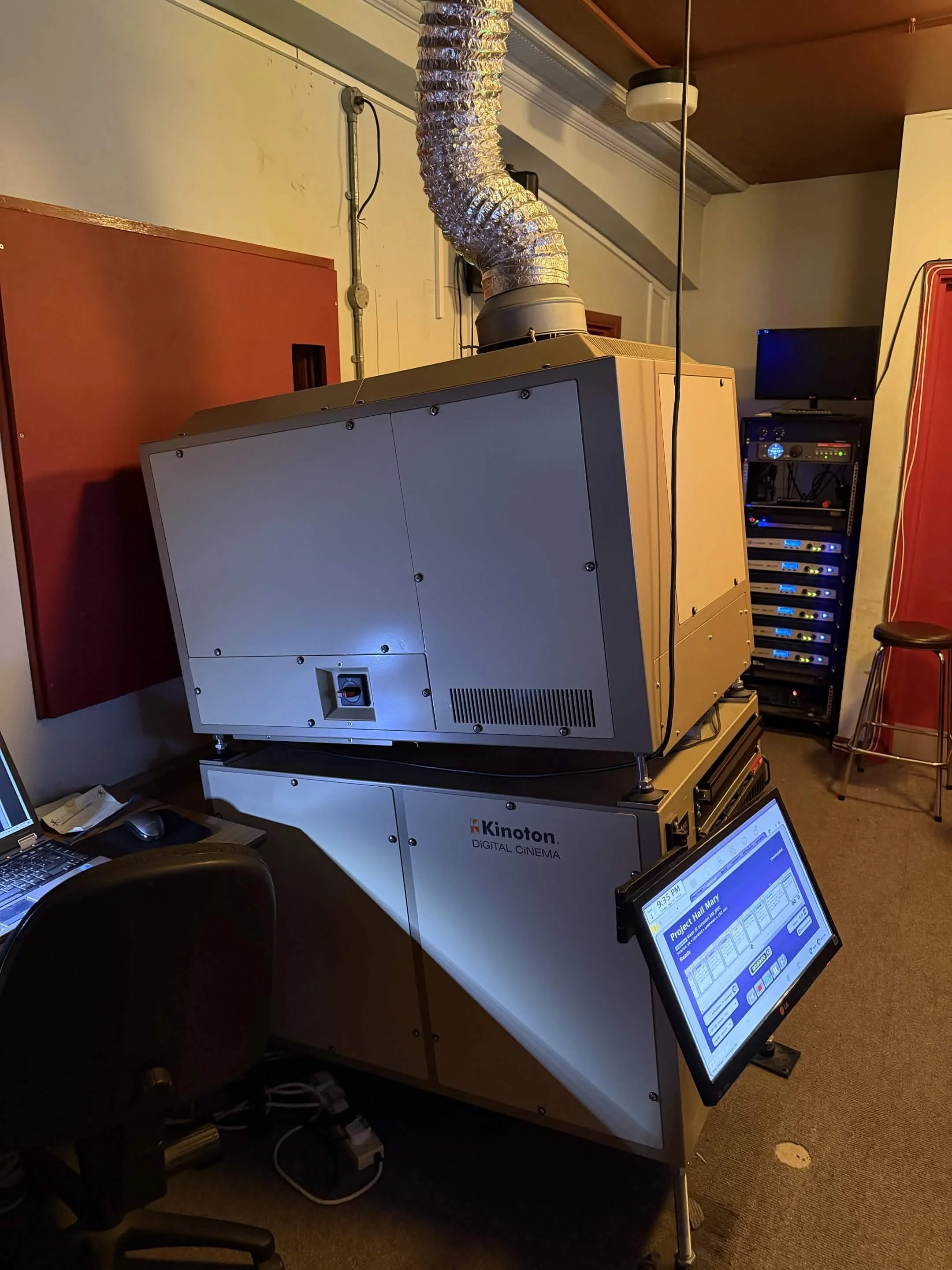 A large digital cinema projector labeled 'Kinoton Digital Cinema' positioned on a table, with a computer monitor and a control touchscreen nearby, in a room with listening and control equipment, chairs, and red and beige walls.