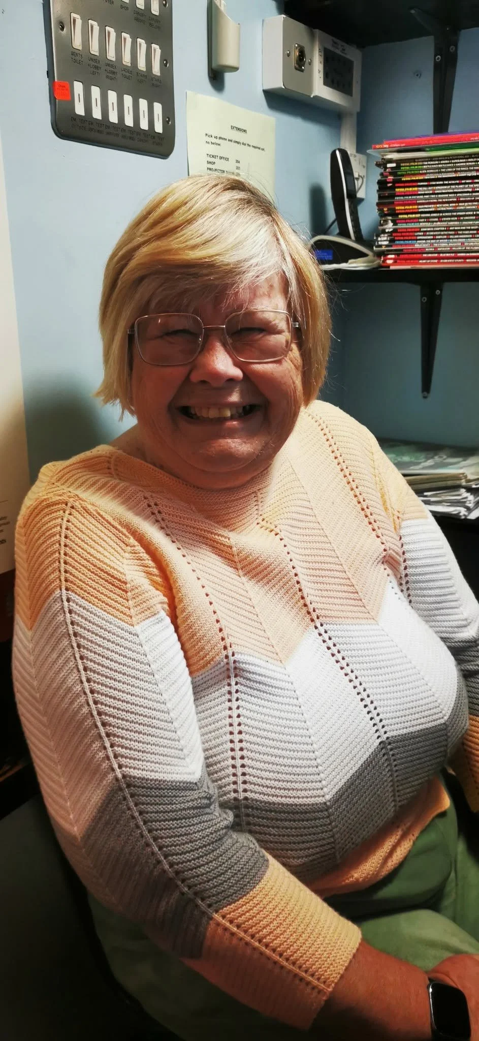 An elderly woman with blonde hair and glasses smiling while sitting in an office. She is wearing a colorful, striped sweater and appears happy.
