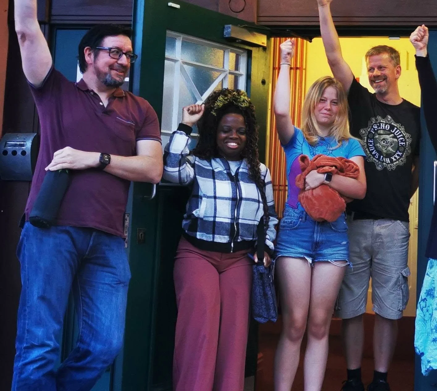 Four people standing in a row inside a restaurant or bar, smiling and raising their arms, celebrating, with colorful walls and a door behind them.
