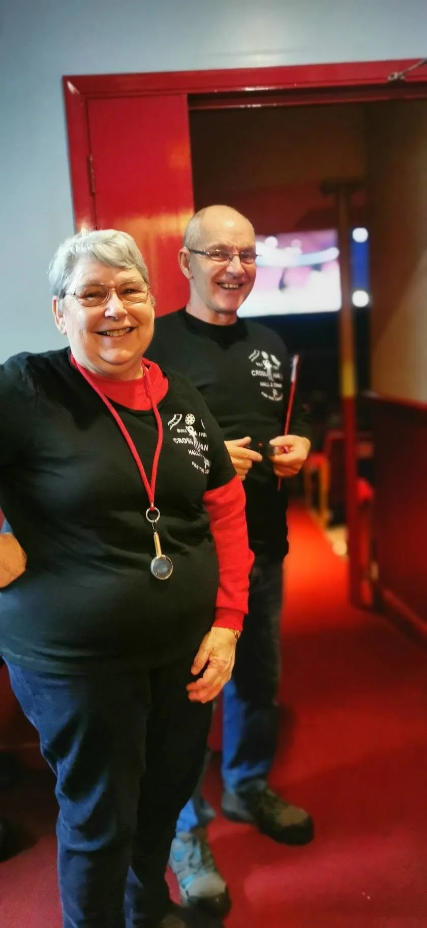 Two smiling people, an elderly woman with short gray hair and glasses, wearing a black shirt with red sleeves, and an elderly man with glasses, wearing a black shirt, standing in a red-themed room with a door open behind them.