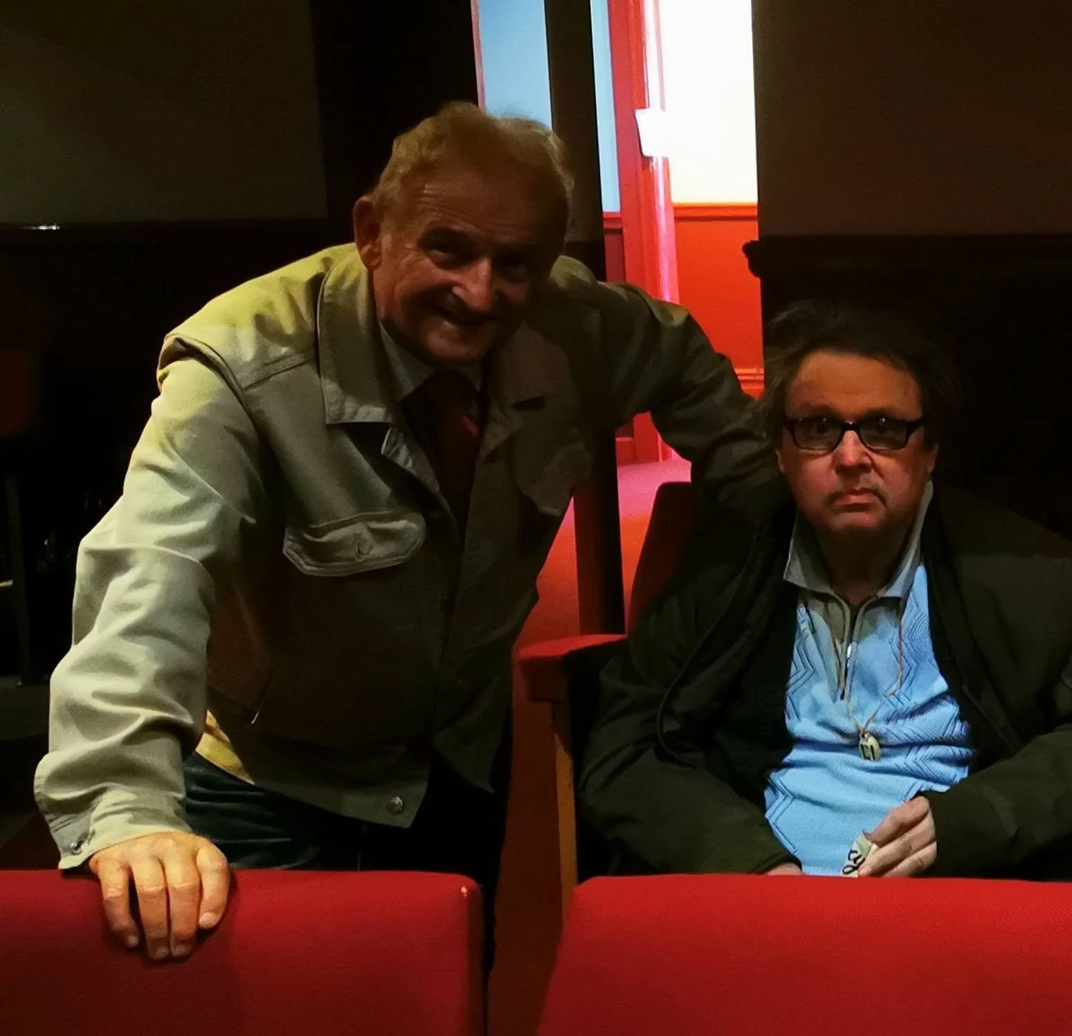 Two men, one standing and one sitting, inside a dimly lit room with red walls and furniture. The standing man has blonde hair and is smiling, wearing a gray jacket and shirt. The seated man has dark hair, glasses, and a serious expression, wearing a 