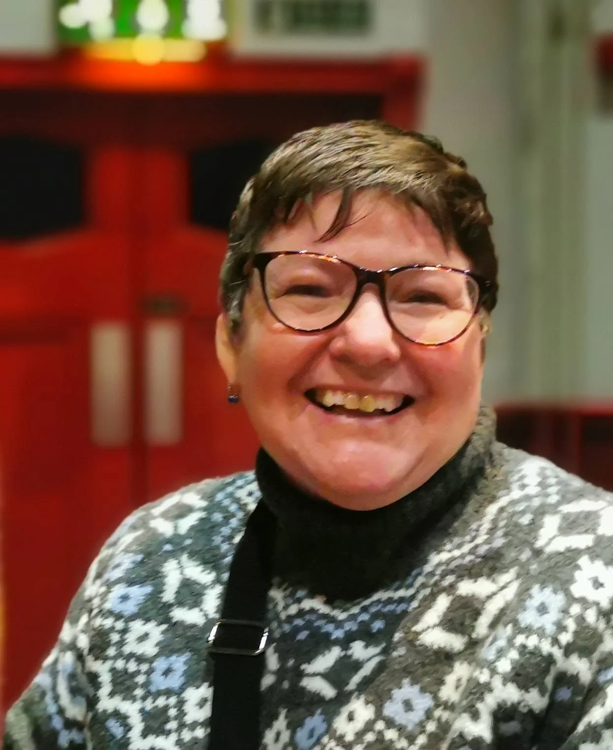 A smiling woman with short brown hair, glasses, and earrings, wearing a patterned sweater and black turtleneck, in a warmly lit room.