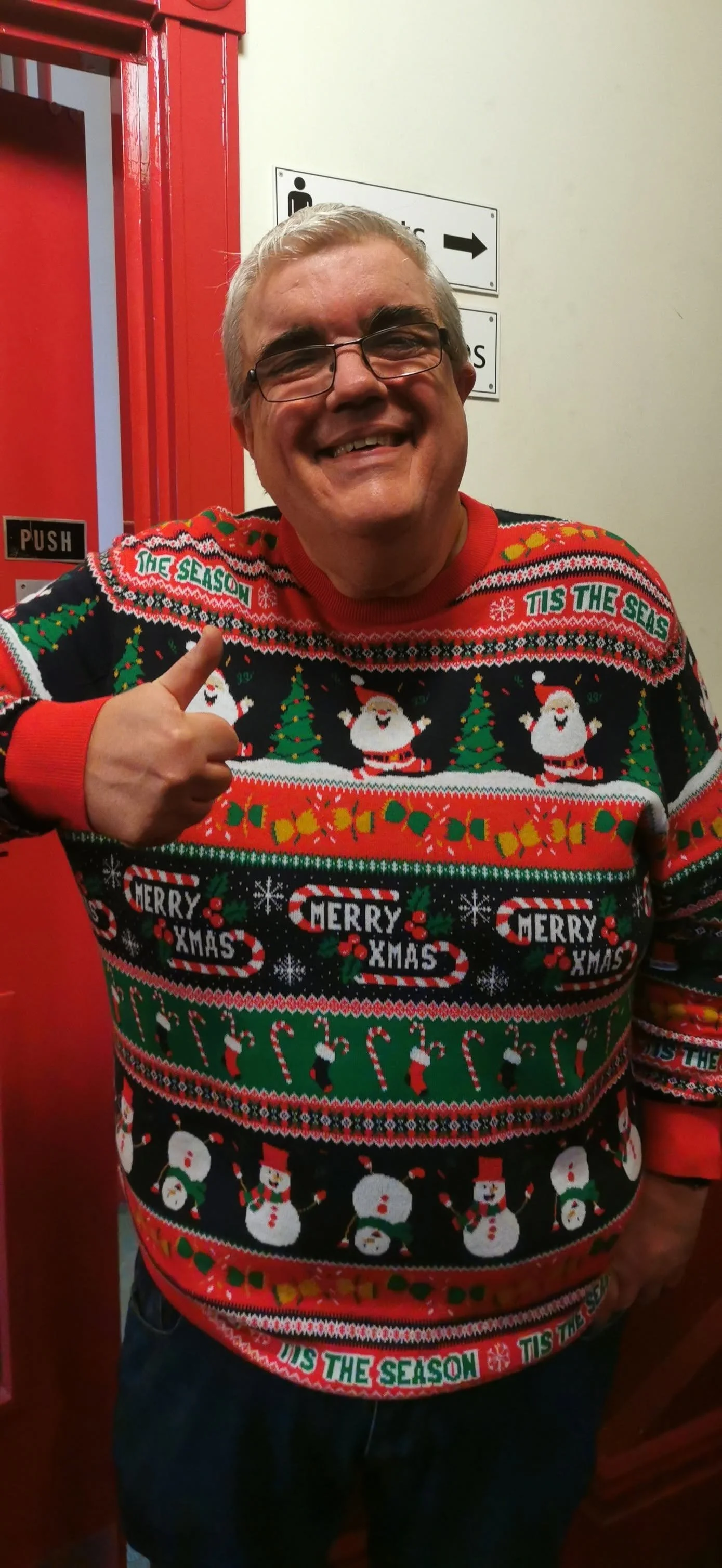 Man wearing a colorful Christmas sweater with snowmen, Santa, Christmas trees, candy canes, and holiday messages, smiling and giving a thumbs up.