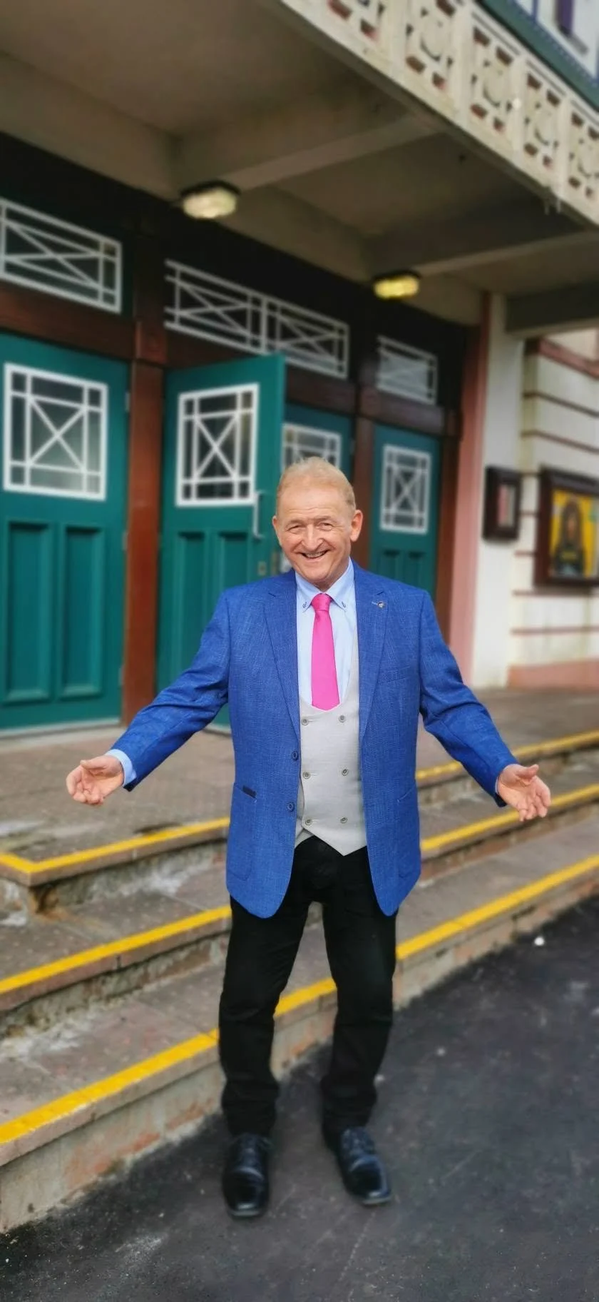 A man in a blue suit and pink tie standing outside, smiling with arms outstretched in front of teal doors and steps.