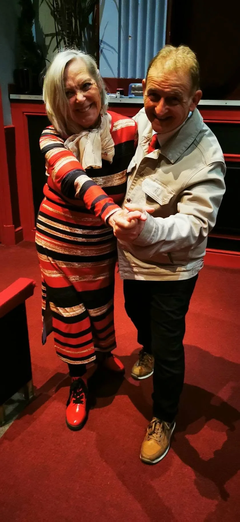 A smiling woman in a colorful striped dress and a man in a light jacket dancing together indoors.