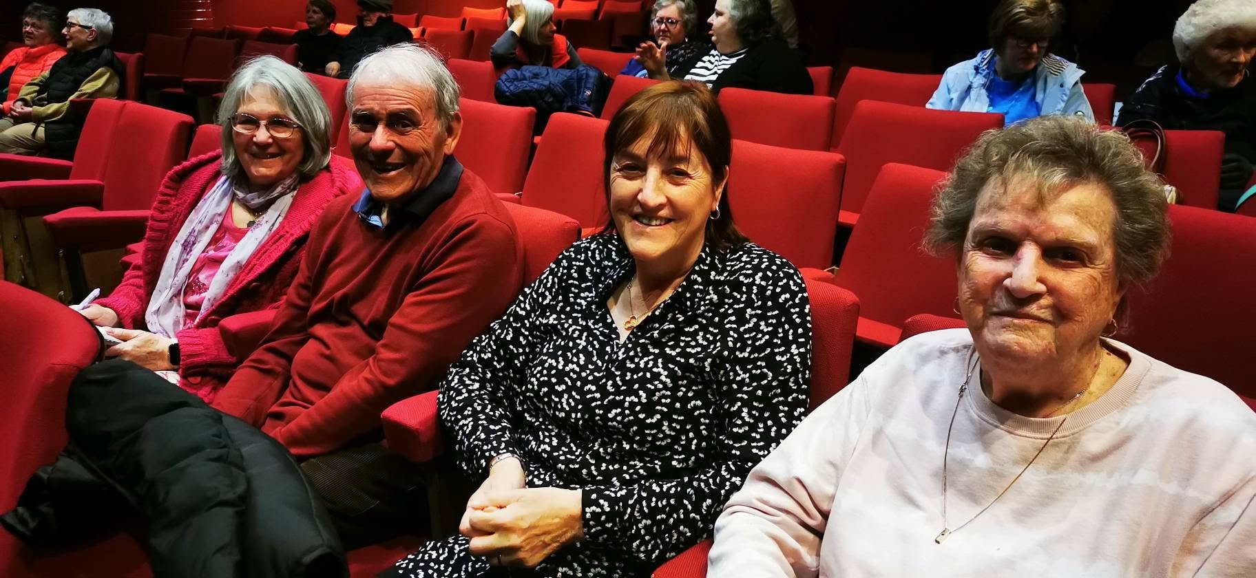Four elderly people sitting in red theater seats, smiling at the camera, with other people in the background.