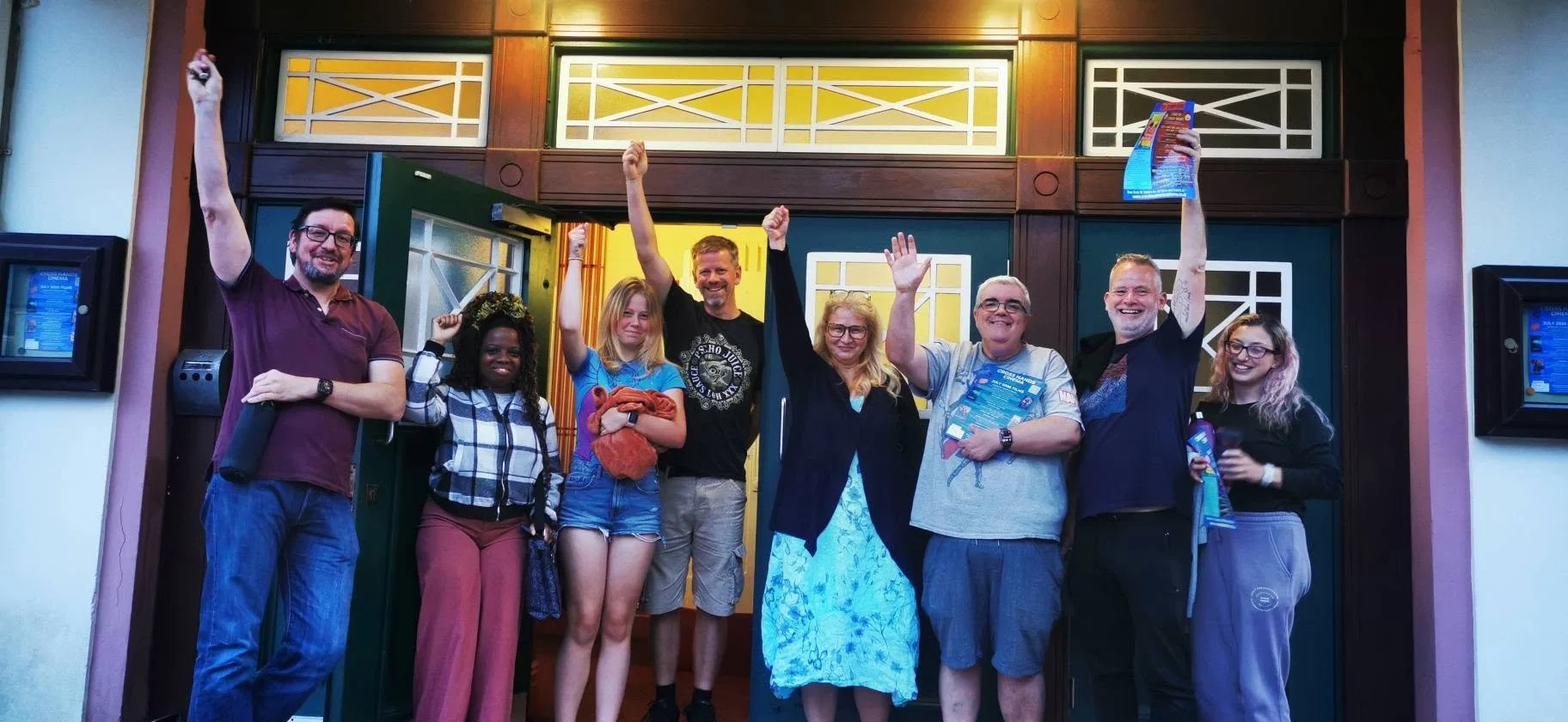 Group of nine people standing in front of a building, celebrating with raised hands, some holding pamphlets or snacks.