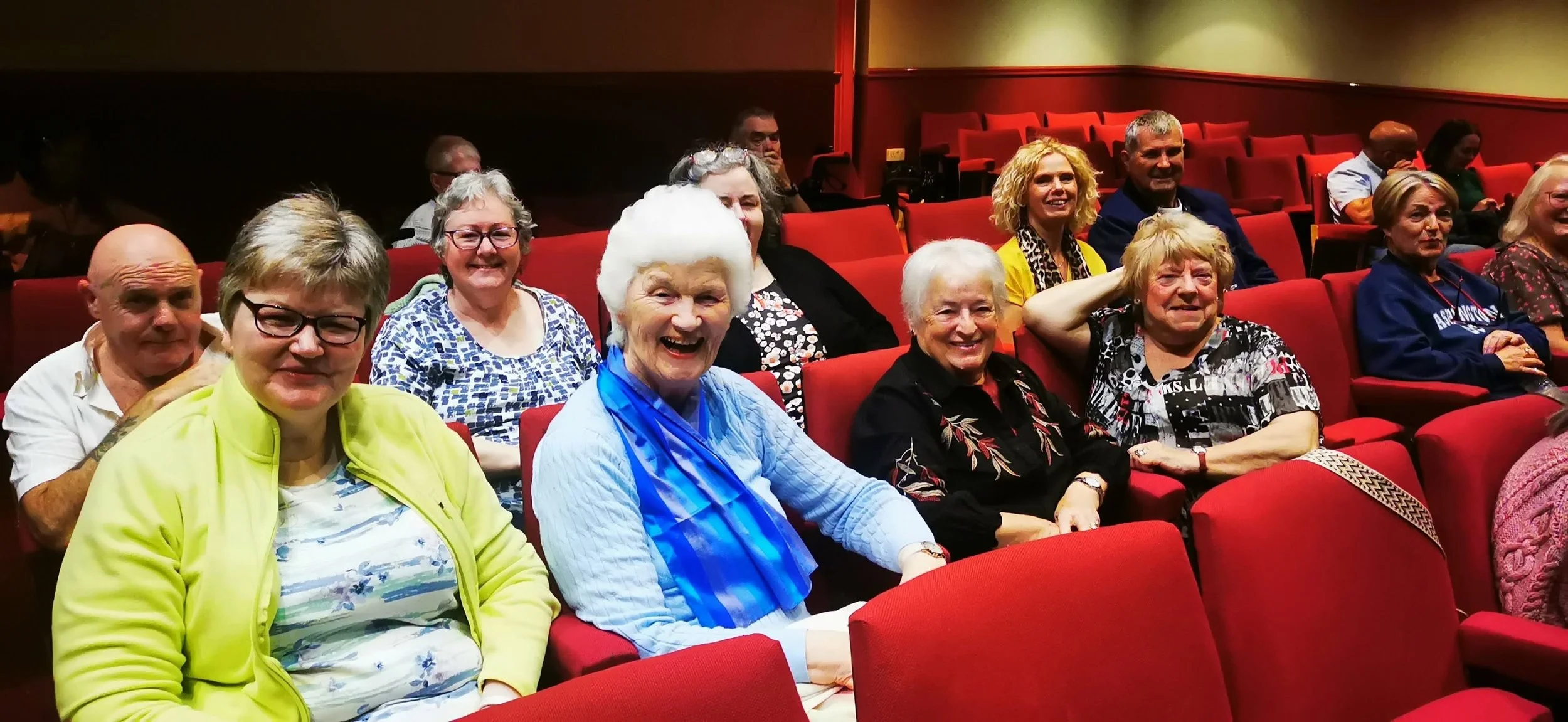 A group of elderly and middle-aged people sitting in a theater with red seats, smiling and looking at the camera.