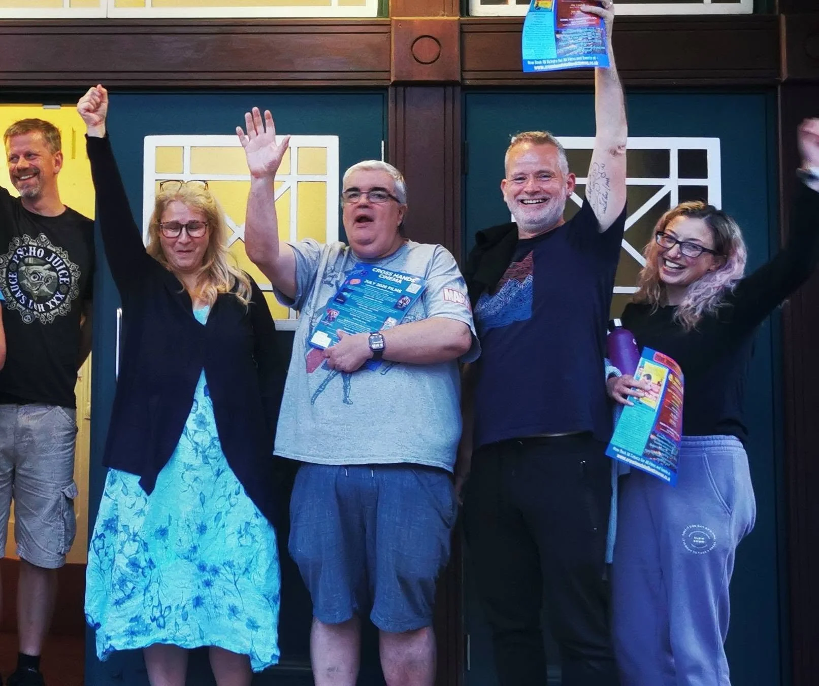Group of five people smiling and raising their hands, holding flyers, standing in front of a wooden wall with windows.
