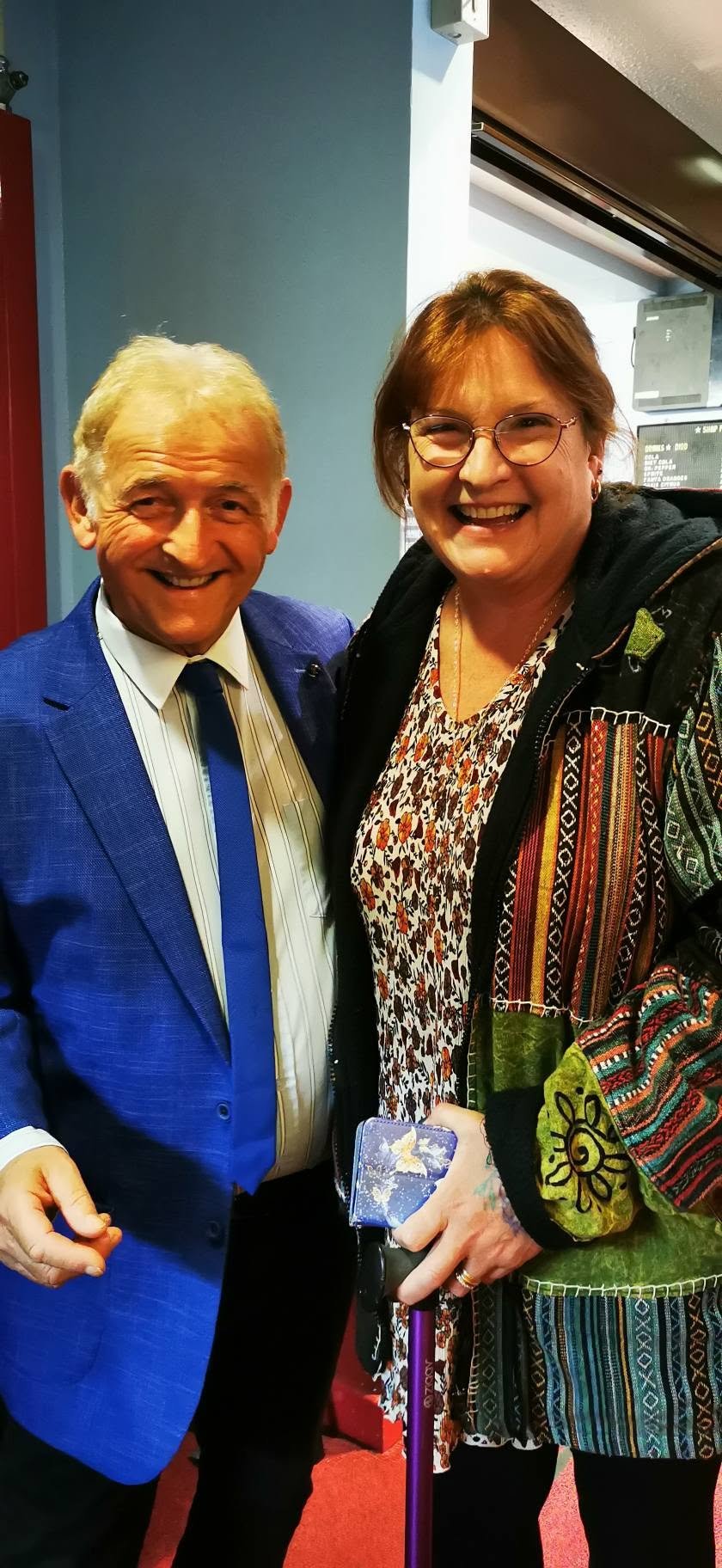 Two smiling women standing close together in an indoor setting, one wearing a bright blue blazer and the other wearing glasses, a patterned shirt, and a colorful jacket. The woman on the right is holding a cane with a decorative top.