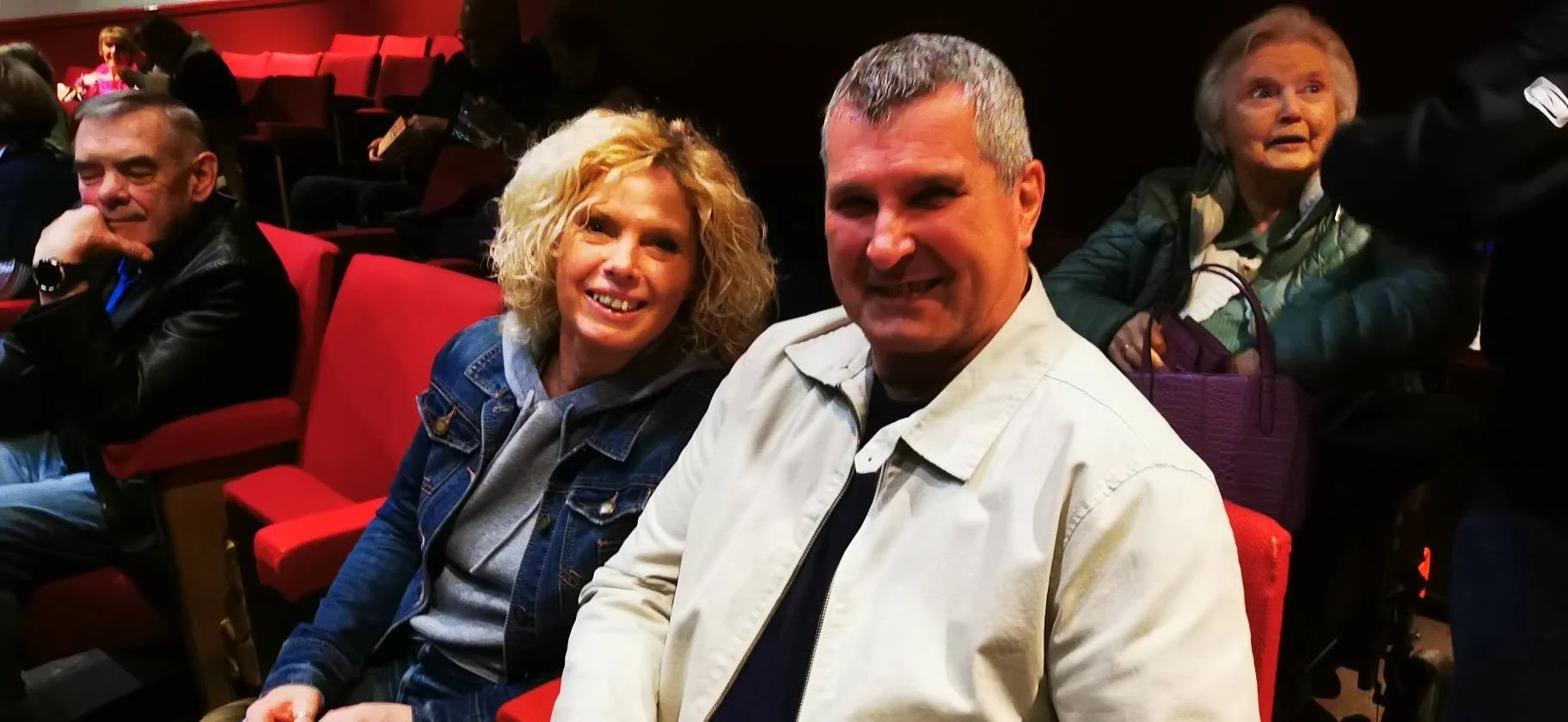 Group of people sitting in red theater seats, smiling and enjoying a show or event, with a woman and man in the foreground and others in the background.