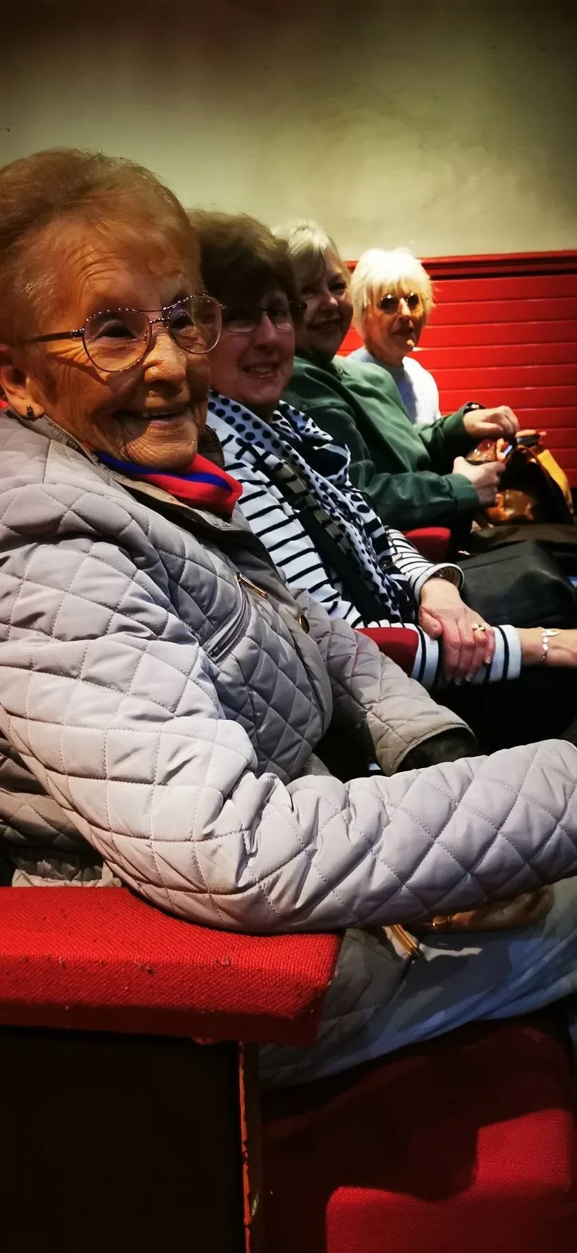 Four elderly women sitting in a row in a theater, smiling and looking at the camera. They are wearing casual jackets and scarves, with red theater seats behind them.