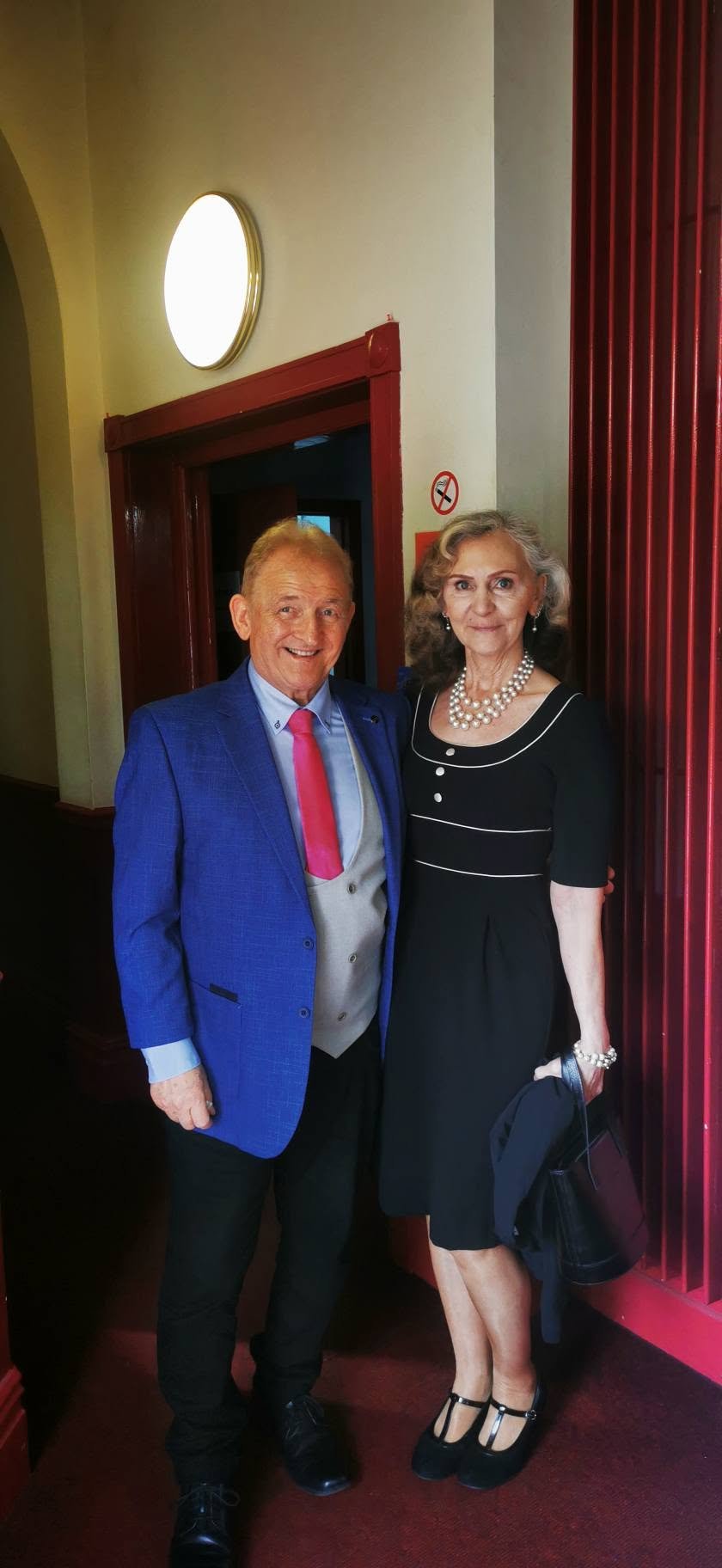 A man and woman dressed formally, standing side by side in a restaurant or event venue, smiling at the camera.