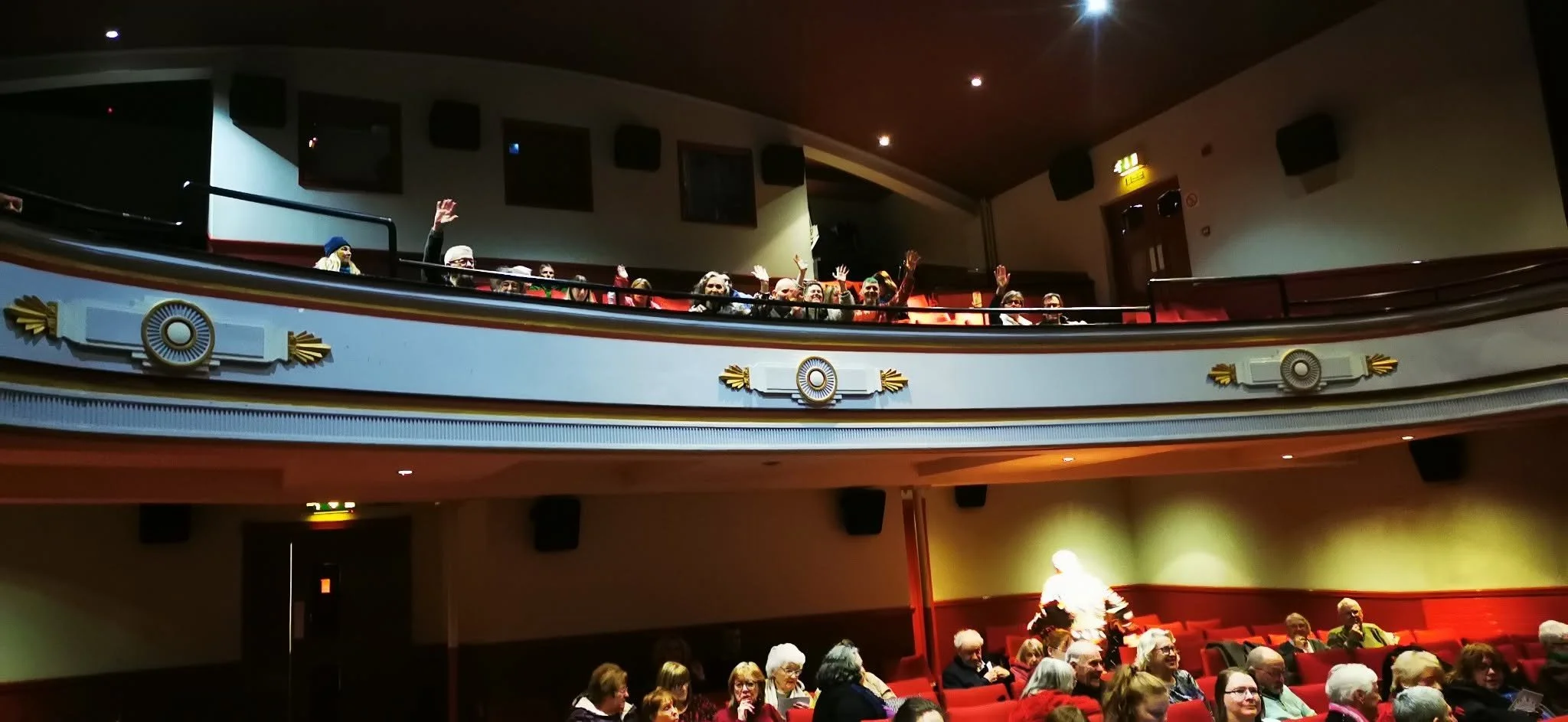 Audience in theater balcony waving at the camera.
