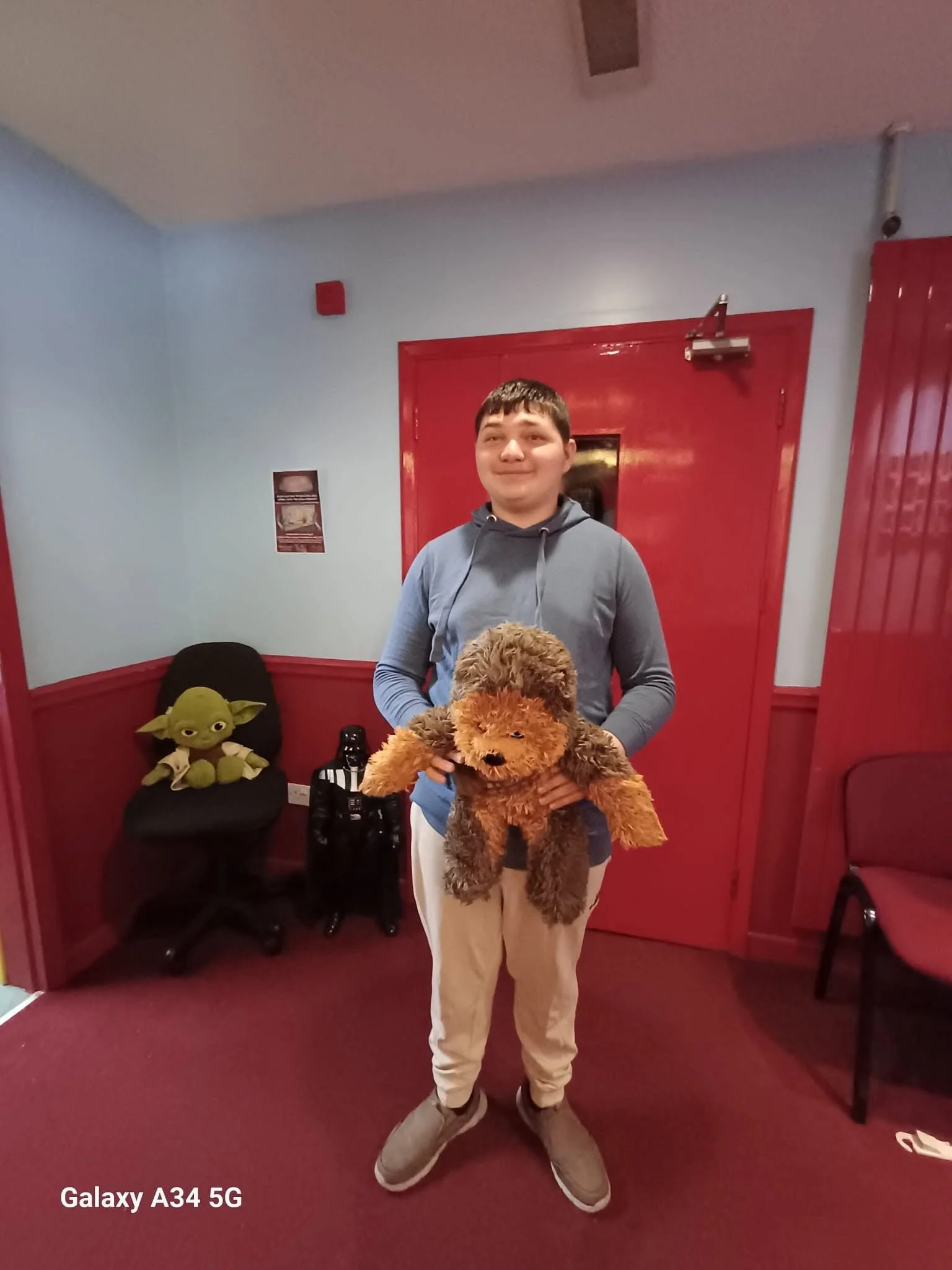 A young person standing in a room with red and blue walls, holding a large brown stuffed dog toy. There is a Yoda plush toy on a black chair to the left and some bottles on the floor nearby. The person is wearing beige pants, a gray hoodie, and sneak