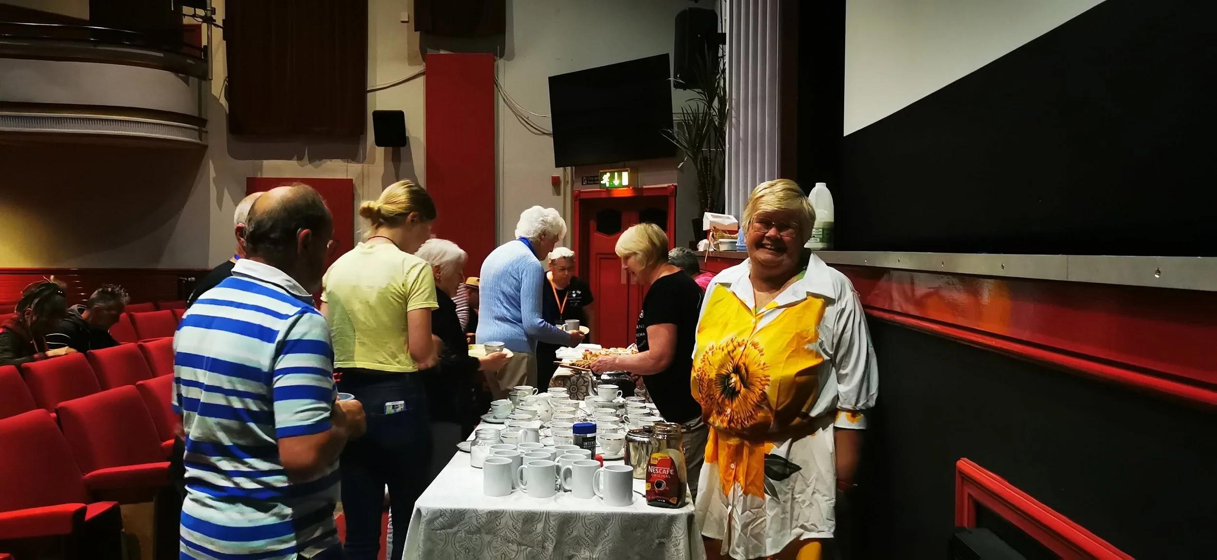 People gathered around a table with coffee cups and jars, serving or enjoying coffee, in a room with red chairs and a large black screen. A woman in a white shirt with a sunflower design is smiling at the camera.