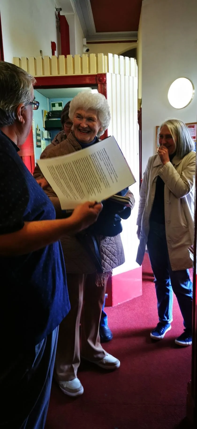 A group of four people standing inside a building, engaged in a conversation. The woman in the center is smiling and holding a booklet. The people are casually dressed, and there is a red carpet on the floor with soft lighting.