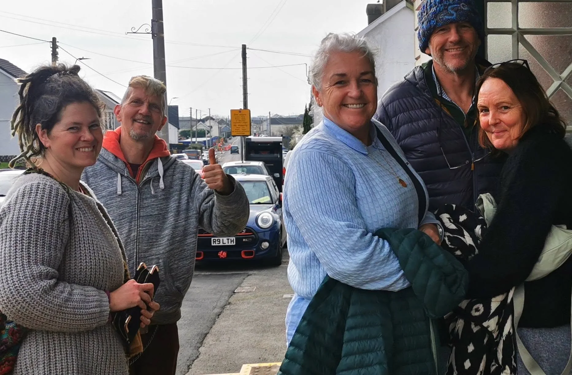 Group of five smiling adults standing on a street with cars in the background, some with jackets and casual clothing.