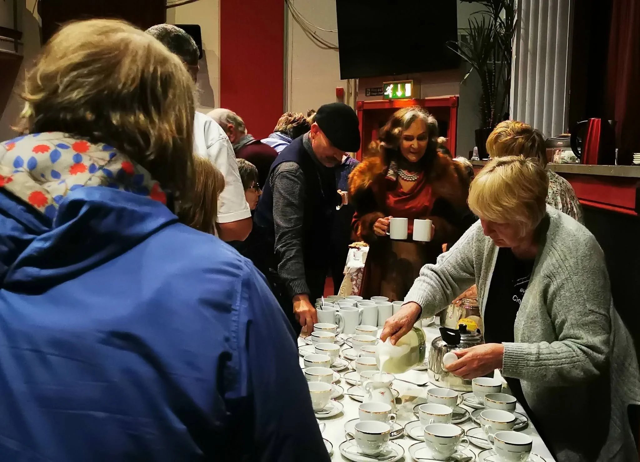 People gathered around a table with multiple white teacups, pouring and serving drinks at an indoor event.