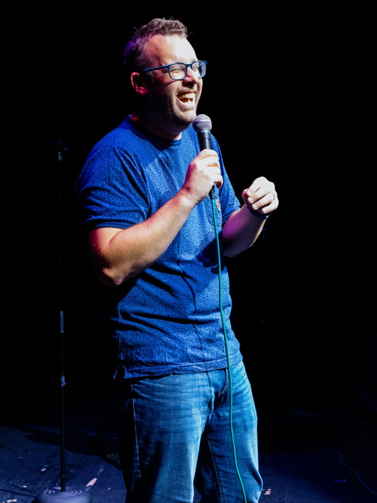 A man with glasses and facial hair, wearing a blue shirt, holding a microphone, smiling while performing on a dark stage.