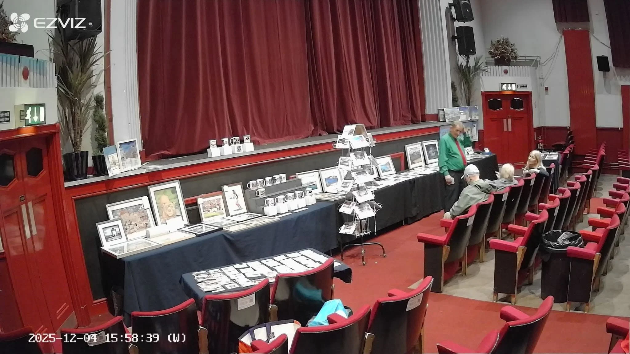 Indoor event space with a stage covered by red curtains. Tables displaying framed pictures, mugs, and other items are set up along the wall. Several people are seated in red chairs, and a man in a green shirt is standing near the tables. The room has