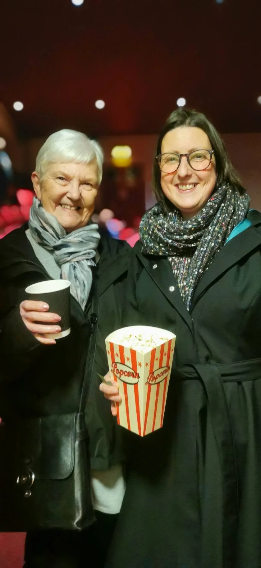 Two smiling women holding popcorn and a drink in a movie theater. The older woman has short white hair, and the younger woman has shoulder-length brown hair and glasses. They are wearing dark coats and scarves, with a dark background and blurred ligh