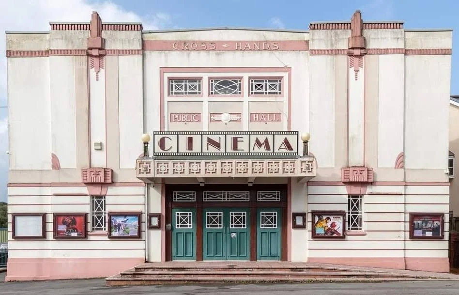 The facade of an art deco style public hall with a cinema entrance, featuring a sign that reads 'CINEMA' and decorative elements in pink and white, including posters and lights.