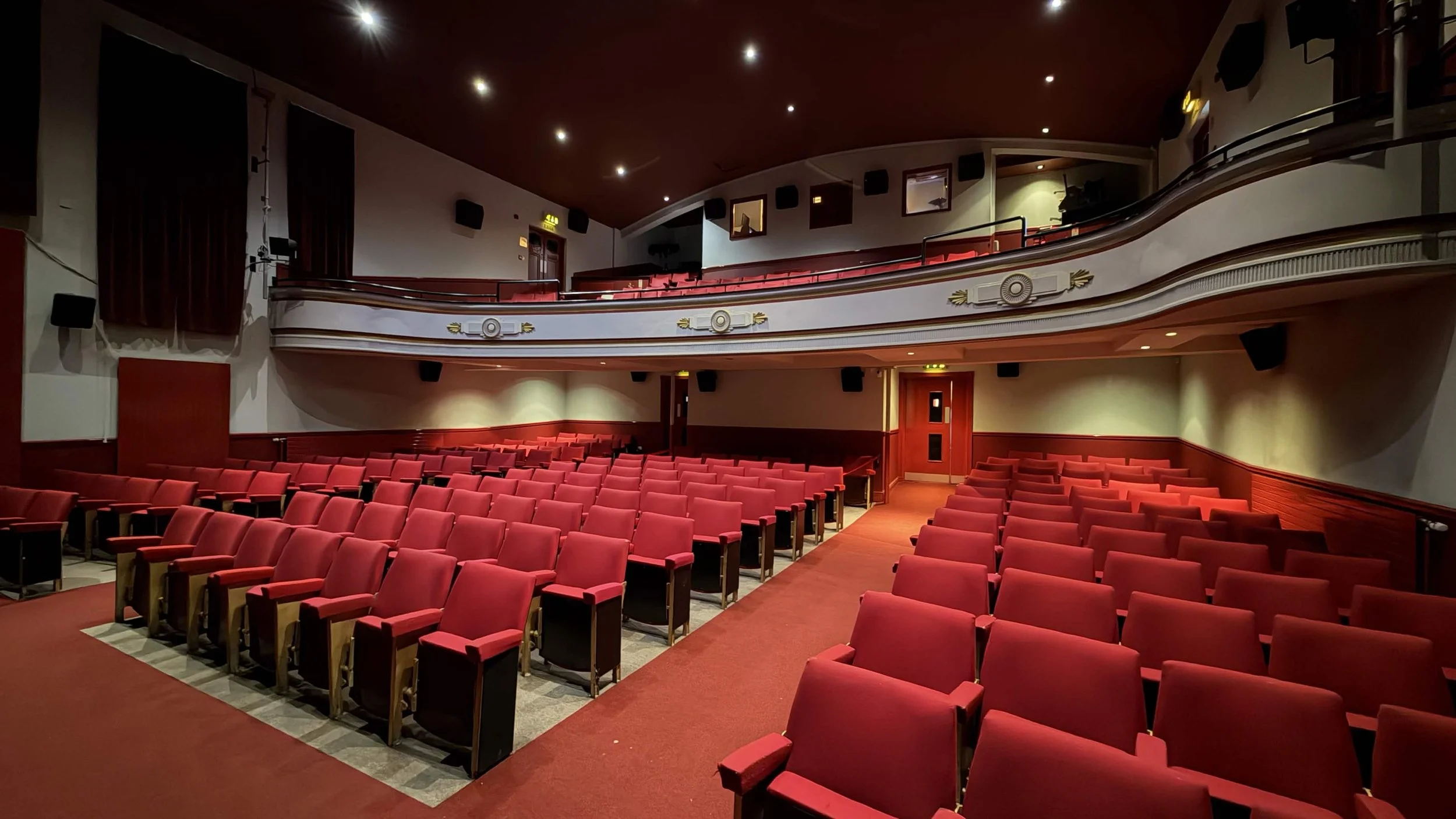 Interior of an empty theater with red upholstered seats, balconies, and stage area, dimly lit with ceiling spotlights.