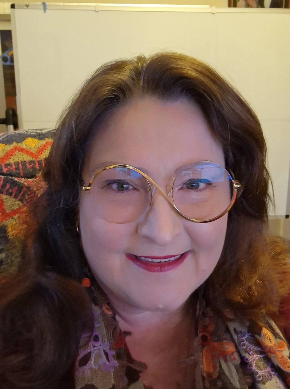 A woman with shoulder-length brown hair, wearing large round glasses with a gold frame, smiling at the camera. She is seated on a patterned cushion with a background that includes a white door and a cabinet.
