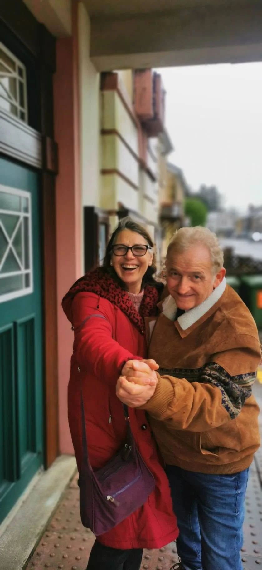 A smiling woman in glasses and a red jacket dancing with a smiling man in a tan jacket on a sidewalk outside a house.