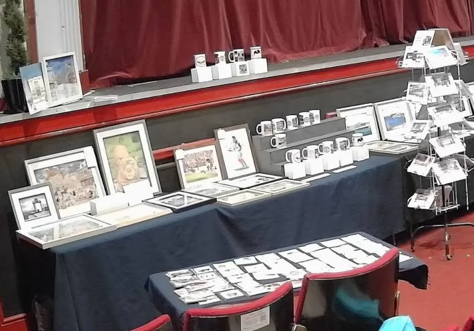 Display table with framed pictures, mugs, postcards, and other small items in a store with a red curtain backdrop.