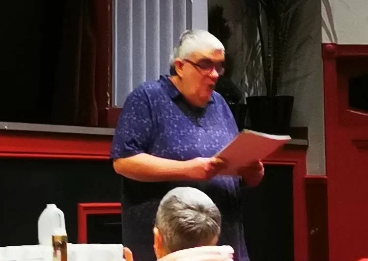An elderly woman with glasses and short white hair standing and reading from a paper in a room with red and black decor, with a person sitting at a table in front of her.