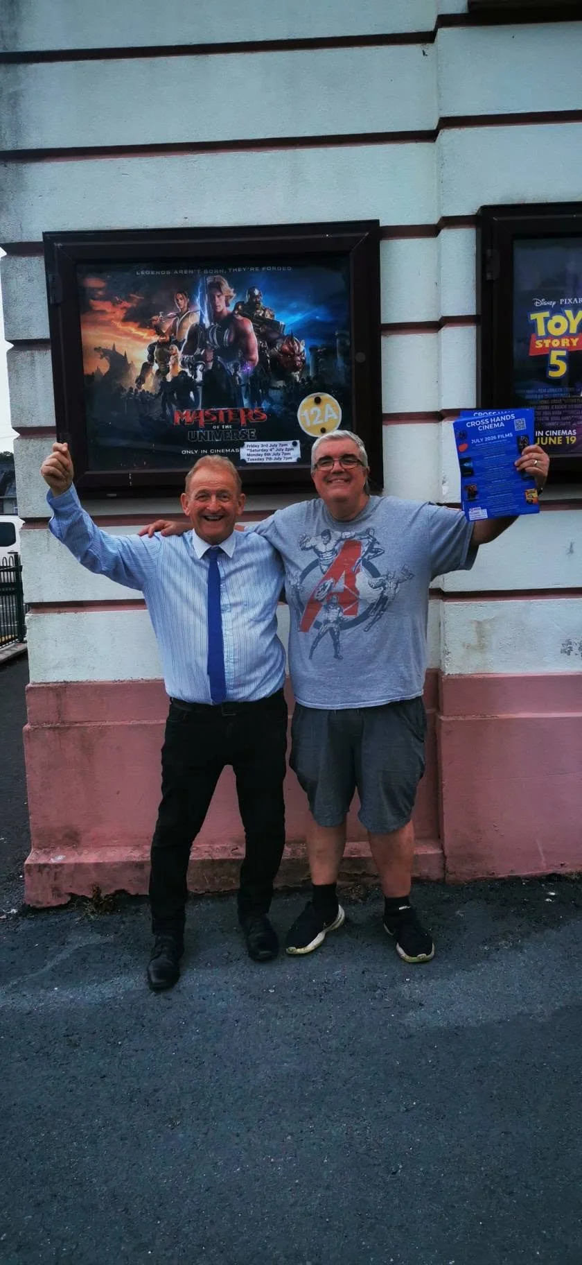 Two men standing outside a movie theater, smiling and celebrating. One man is wearing a shirt and tie, and the other is wearing a gray t-shirt and shorts. They are standing in front of a poster for the movie "Masters of the Universe" and holding a pa