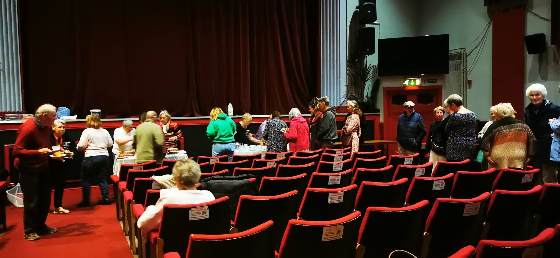 People standing in line at a buffet table inside a theater with red chairs, a large screen, and dark curtains.