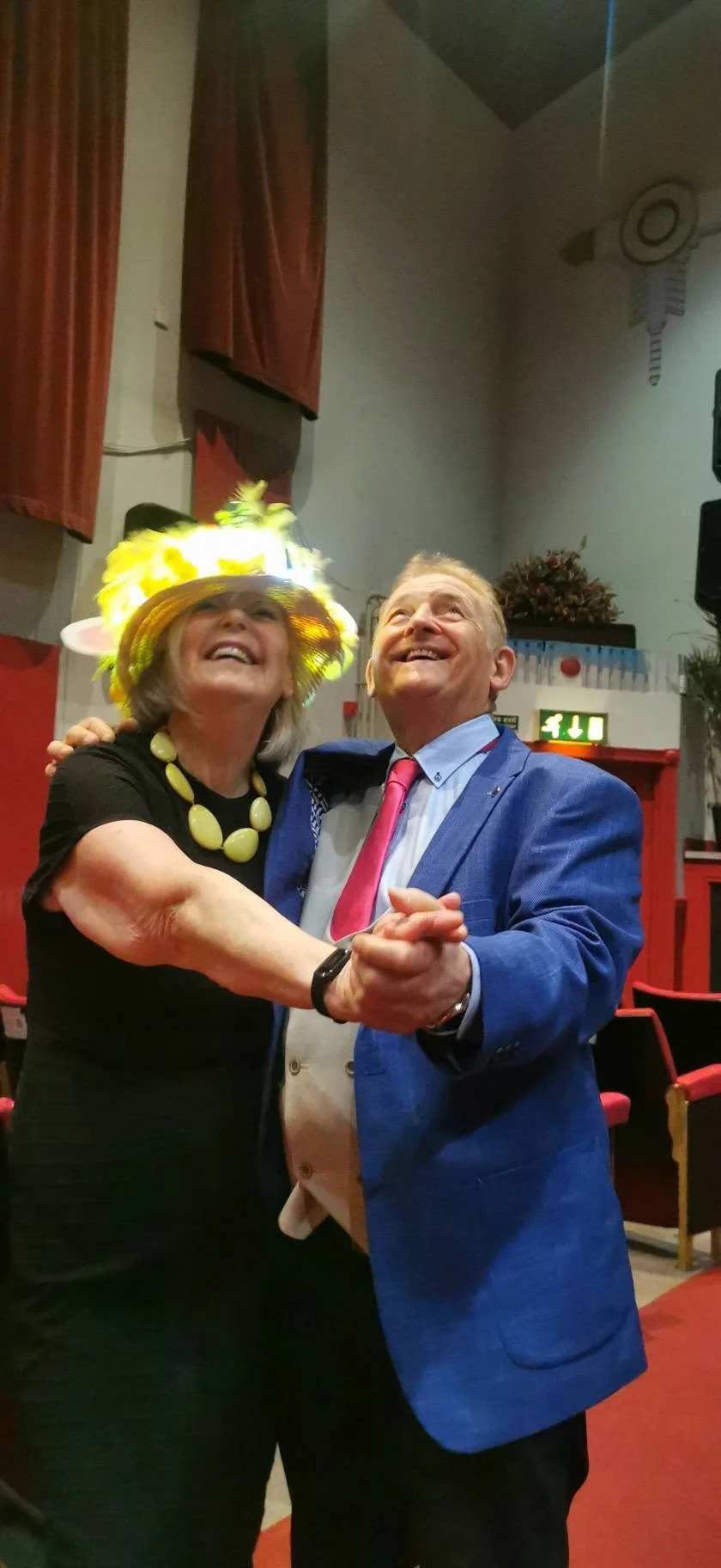 Senior woman and man dancing together indoors, woman wearing bright hat and necklace, man in blue suit with pink tie, both smiling.